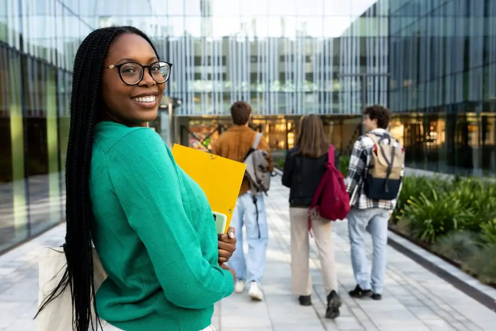 Smiling student holding a folder outside a university building as classmates walk ahead