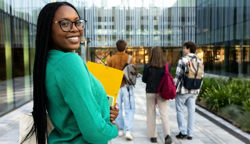 Smiling student holding a folder outside a university building as classmates walk ahead
